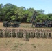 A large group of military personnel from what appears to be two different nations pose together in a field for a group photograph. Behind them are two military rocket/missile launcher systems mounted on heavy tactical trucks. The setting appears to be an open training area with trees in the background, likely during a joint military exercise.