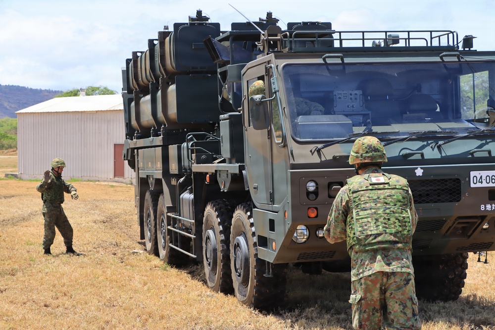 Two soldiers in Japanese Ground Self-Defense Force (JGSDF) camouflage uniforms are directing or guiding a large military truck-mounted missile launcher system. The vehicle appears to be a Type 12 Surface-to-Ship Missile (SSM) launcher mounted on a heavy tactical truck. The setting appears to be an open field or training area, possibly in Hawaii based on the terrain and vegetation.