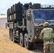 Two soldiers in Japanese Ground Self-Defense Force (JGSDF) camouflage uniforms are directing or guiding a large military truck-mounted missile launcher system. The vehicle appears to be a Type 12 Surface-to-Ship Missile (SSM) launcher mounted on a heavy tactical truck. The setting appears to be an open field or training area, possibly in Hawaii based on the terrain and vegetation.