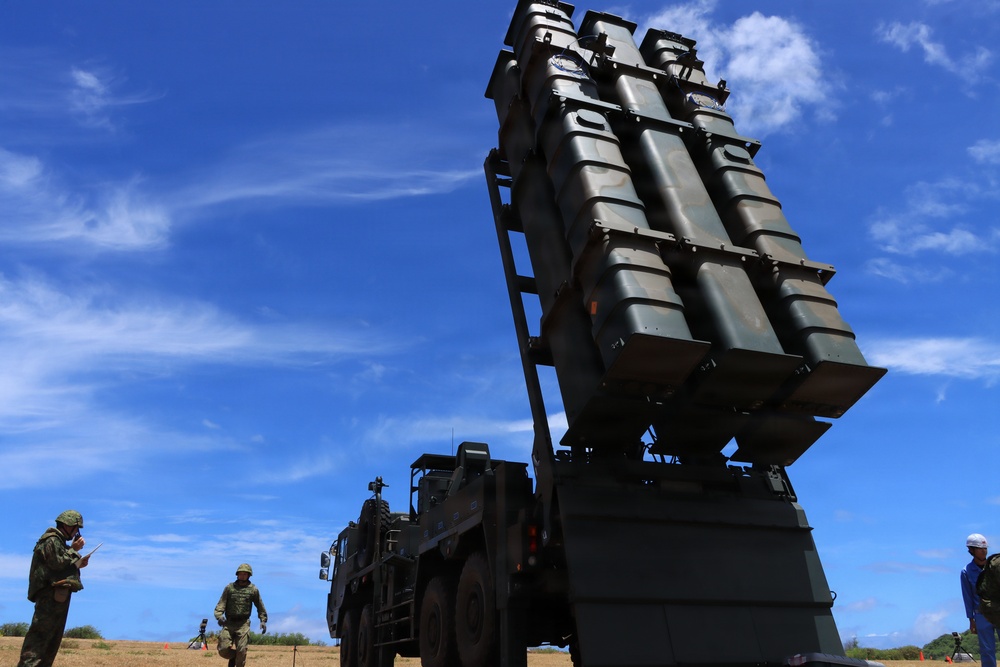 The image shows a large military missile launcher system mounted on a heavy tactical truck, elevated at an angle against a blue sky. Several military personnel in camouflage uniforms are visible nearby, along with a civilian in a white hard hat. The setting appears to be an open field or coastal area, likely during a military exercise or demonstration.