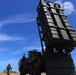 The image shows a large military missile launcher system mounted on a heavy tactical truck, elevated at an angle against a blue sky. Several military personnel in camouflage uniforms are visible nearby, along with a civilian in a white hard hat. The setting appears to be an open field or coastal area, likely during a military exercise or demonstration.