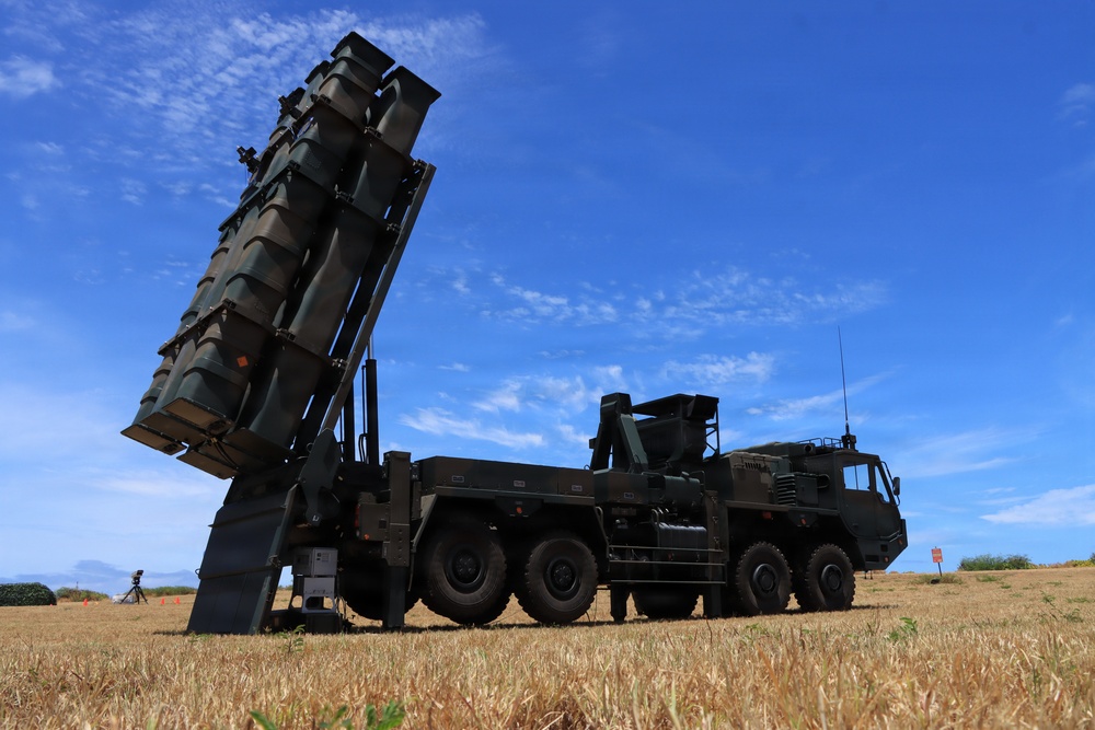 The image shows a large military mobile missile launcher system mounted on a heavy multi-axle truck, deployed in an open grassy field. The launcher is elevated at an angle, with multiple missile canisters visible in a vertical array. The setting appears to be an outdoor military exercise or static display under clear blue skies.