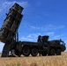 The image shows a large military mobile missile launcher system mounted on a heavy multi-axle truck, deployed in an open grassy field. The launcher is elevated at an angle, with multiple missile canisters visible in a vertical array. The setting appears to be an outdoor military exercise or static display under clear blue skies.