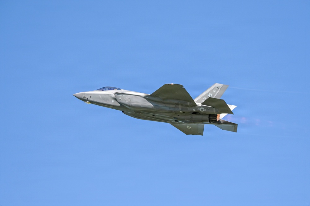 A single-engine stealth fighter jet, identifiable as an F-35A Lightning II, is captured in flight against a clear blue sky. The aircraft is flying in a slightly banked orientation with its afterburner visibly engaged, producing a bright orange-white flame at the exhaust nozzle. Tail markings indicate 'WI', suggesting association with a Wisconsin Air National Guard unit.