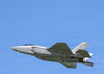 A single-engine stealth fighter jet, identifiable as an F-35A Lightning II, is captured in flight against a clear blue sky. The aircraft is flying in a slightly banked orientation with its afterburner visibly engaged, producing a bright orange-white flame at the exhaust nozzle. Tail markings indicate 'WI', suggesting association with a Wisconsin Air National Guard unit.