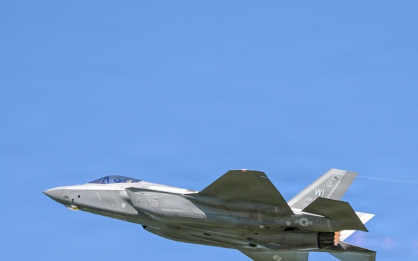 A single-engine stealth fighter jet, identifiable as an F-35A Lightning II, is captured in flight against a clear blue sky. The aircraft is flying in a slightly banked orientation with its afterburner visibly engaged, producing a bright orange-white flame at the exhaust nozzle. Tail markings indicate 'WI', suggesting association with a Wisconsin Air National Guard unit.