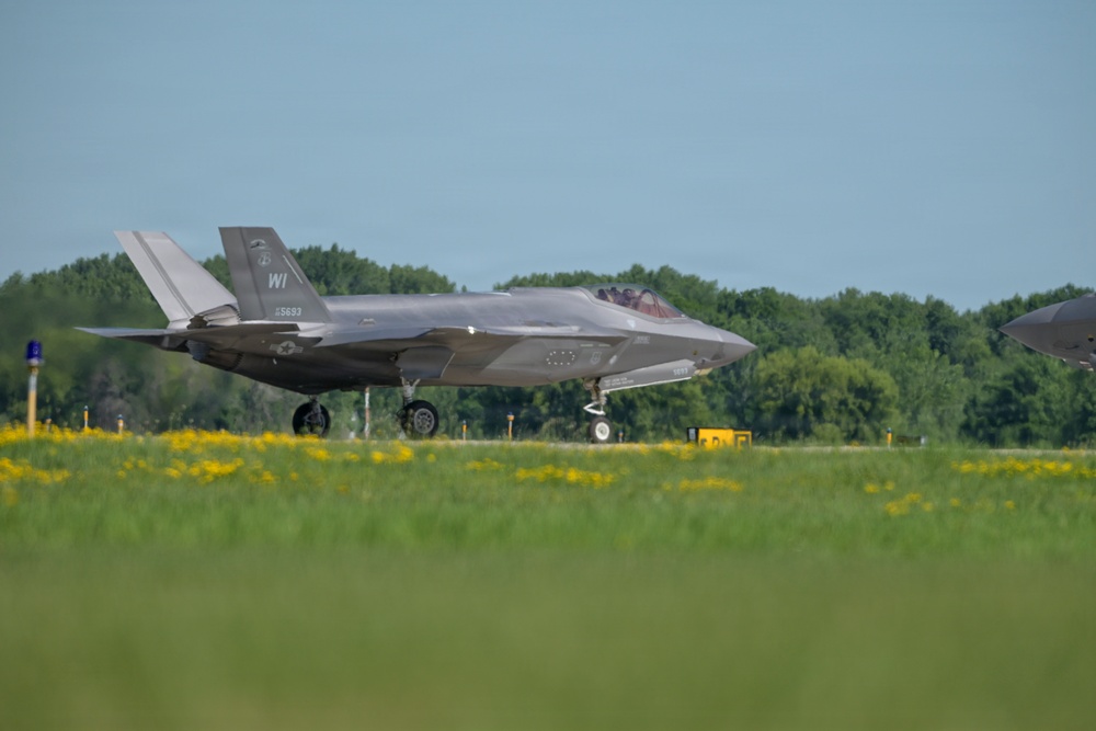 An F-35A Lightning II stealth fighter jet is taxiing on a runway apron, displaying 'WI' tail codes indicating Wisconsin Air National Guard assignment. The aircraft bears the serial number 15-5693 on its tail fin and appears to be in standard gray low-visibility paint scheme. A second aircraft is partially visible on the far right edge of the frame.
