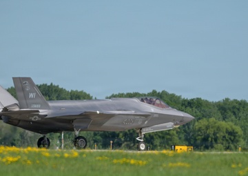 An F-35A Lightning II stealth fighter jet is taxiing on a runway apron, displaying 'WI' tail codes indicating Wisconsin Air National Guard assignment. The aircraft bears the serial number 15-5693 on its tail fin and appears to be in standard gray low-visibility paint scheme. A second aircraft is partially visible on the far right edge of the frame.