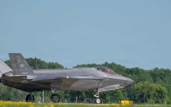 An F-35A Lightning II stealth fighter jet is taxiing on a runway apron, displaying 'WI' tail codes indicating Wisconsin Air National Guard assignment. The aircraft bears the serial number 15-5693 on its tail fin and appears to be in standard gray low-visibility paint scheme. A second aircraft is partially visible on the far right edge of the frame.
