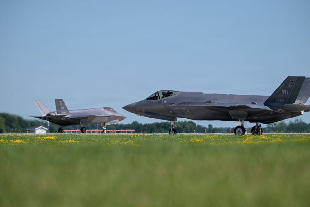 Two F-35A Lightning II stealth fighter jets are taxiing on a runway at what appears to be a military airfield. Both aircraft display 'WI' tail codes, indicating they belong to the Wisconsin Air National Guard. A ground crew member is visible near the foreground aircraft, and the setting features green grass with yellow wildflowers in the foreground.