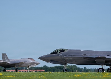 Two F-35A Lightning II stealth fighter jets are taxiing on a runway at what appears to be a military airfield. Both aircraft display 'WI' tail codes, indicating they belong to the Wisconsin Air National Guard. A ground crew member is visible near the foreground aircraft, and the setting features green grass with yellow wildflowers in the foreground.
