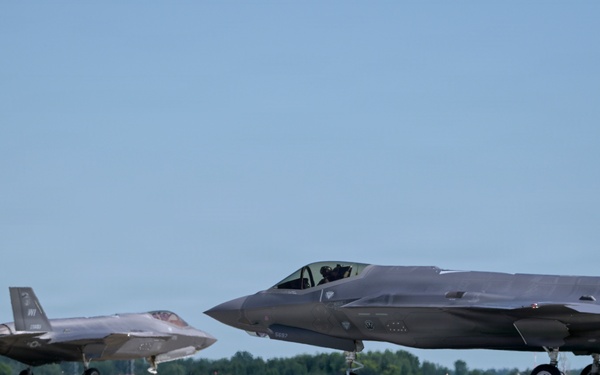 Two F-35A Lightning II stealth fighter jets are taxiing on a runway at what appears to be a military airfield. Both aircraft display 'WI' tail codes, indicating they belong to the Wisconsin Air National Guard. A ground crew member is visible near the foreground aircraft, and the setting features green grass with yellow wildflowers in the foreground.