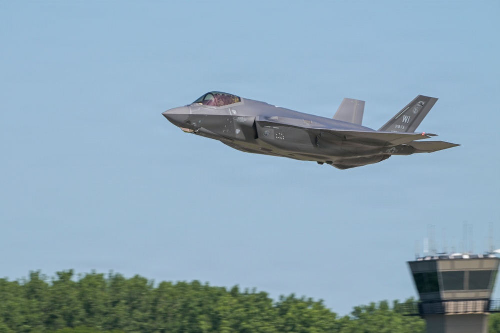 A single-engine stealth fighter jet, identifiable as an F-35A Lightning II, is captured in low-altitude flight near an air traffic control tower. The aircraft bears tail markings 'WI' and '115 FS' (or similar), indicating it belongs to a Wisconsin Air National Guard unit. The image was likely taken at or near a military airfield during a takeoff or low-level pass.