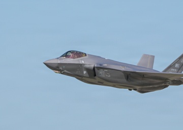 A single-engine stealth fighter jet, identifiable as an F-35A Lightning II, is captured in low-altitude flight near an air traffic control tower. The aircraft bears tail markings 'WI' and '115 FS' (or similar), indicating it belongs to a Wisconsin Air National Guard unit. The image was likely taken at or near a military airfield during a takeoff or low-level pass.