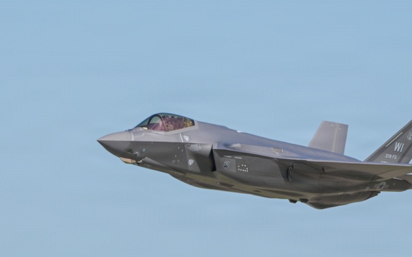 A single-engine stealth fighter jet, identifiable as an F-35A Lightning II, is captured in low-altitude flight near an air traffic control tower. The aircraft bears tail markings 'WI' and '115 FS' (or similar), indicating it belongs to a Wisconsin Air National Guard unit. The image was likely taken at or near a military airfield during a takeoff or low-level pass.