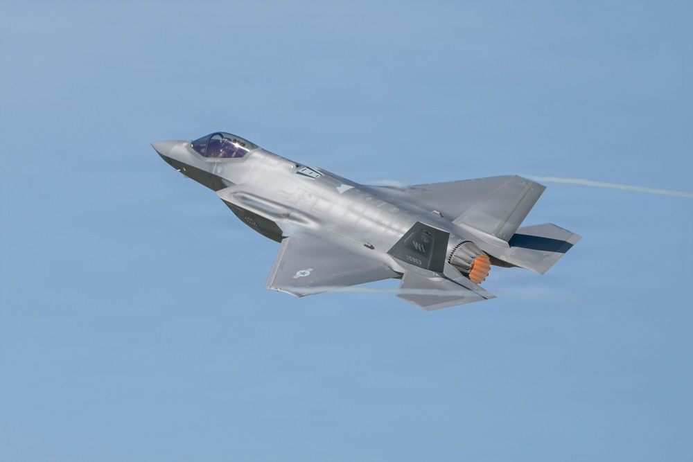 A single-engine stealth multirole fighter jet, consistent with an F-35 Lightning II, is captured in flight against a clear blue sky. The aircraft is in a slight bank, with its afterburner visibly glowing orange at the exhaust nozzle. A faint condensation trail is visible trailing from the wingtip area.