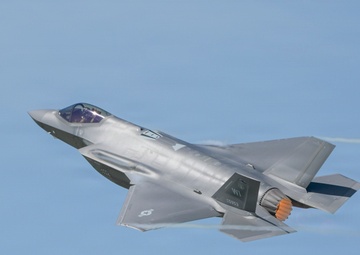 A single-engine stealth multirole fighter jet, consistent with an F-35 Lightning II, is captured in flight against a clear blue sky. The aircraft is in a slight bank, with its afterburner visibly glowing orange at the exhaust nozzle. A faint condensation trail is visible trailing from the wingtip area.