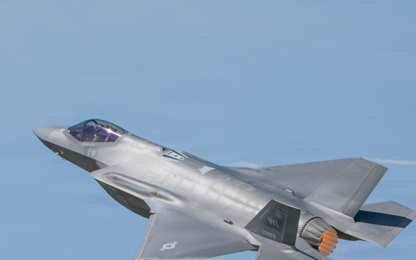A single-engine stealth multirole fighter jet, consistent with an F-35 Lightning II, is captured in flight against a clear blue sky. The aircraft is in a slight bank, with its afterburner visibly glowing orange at the exhaust nozzle. A faint condensation trail is visible trailing from the wingtip area.