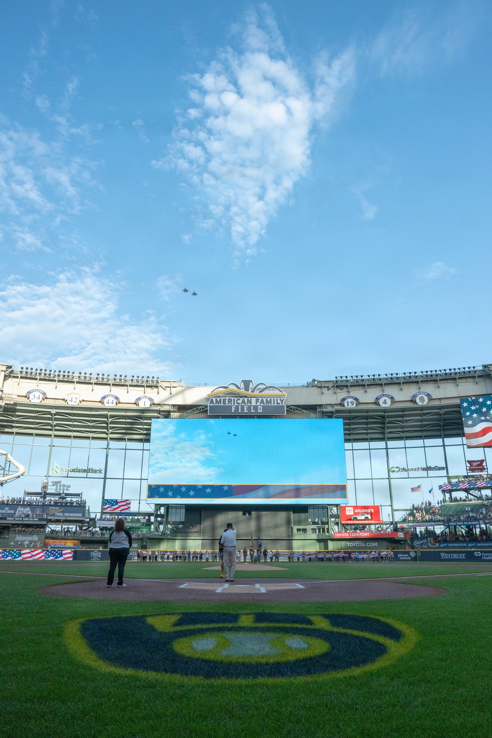 A pre-game ceremony is taking place at American Family Field, home of the Milwaukee Brewers MLB team. Two military aircraft are performing a flyover above the stadium, which is simultaneously displayed on the large center field scoreboard. Two individuals stand near home plate on the field, likely during the national anthem.