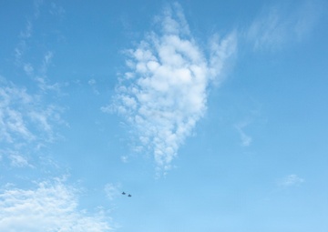 A pre-game ceremony is taking place at American Family Field, home of the Milwaukee Brewers MLB team. Two military aircraft are performing a flyover above the stadium, which is simultaneously displayed on the large center field scoreboard. Two individuals stand near home plate on the field, likely during the national anthem.