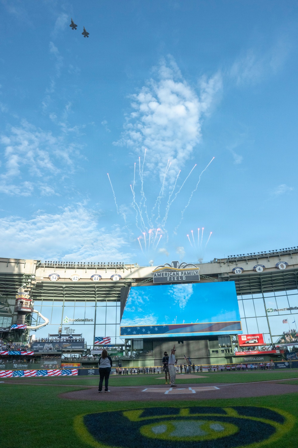 A pre-game ceremony is taking place at American Family Field, home of the Milwaukee Brewers MLB team. Two military jet aircraft are performing a flyover while fireworks are simultaneously launched above the stadium. The scene appears to be a patriotic celebration, likely for a holiday such as Independence Day or Memorial Day.