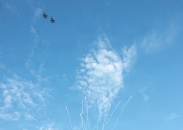 A pre-game ceremony is taking place at American Family Field, home of the Milwaukee Brewers MLB team. Two military jet aircraft are performing a flyover while fireworks are simultaneously launched above the stadium. The scene appears to be a patriotic celebration, likely for a holiday such as Independence Day or Memorial Day.