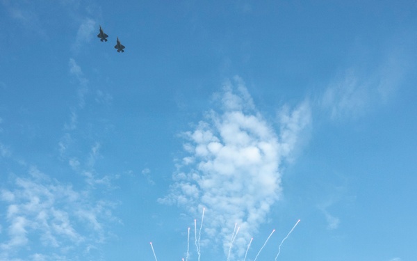 A pre-game ceremony is taking place at American Family Field, home of the Milwaukee Brewers MLB team. Two military jet aircraft are performing a flyover while fireworks are simultaneously launched above the stadium. The scene appears to be a patriotic celebration, likely for a holiday such as Independence Day or Memorial Day.