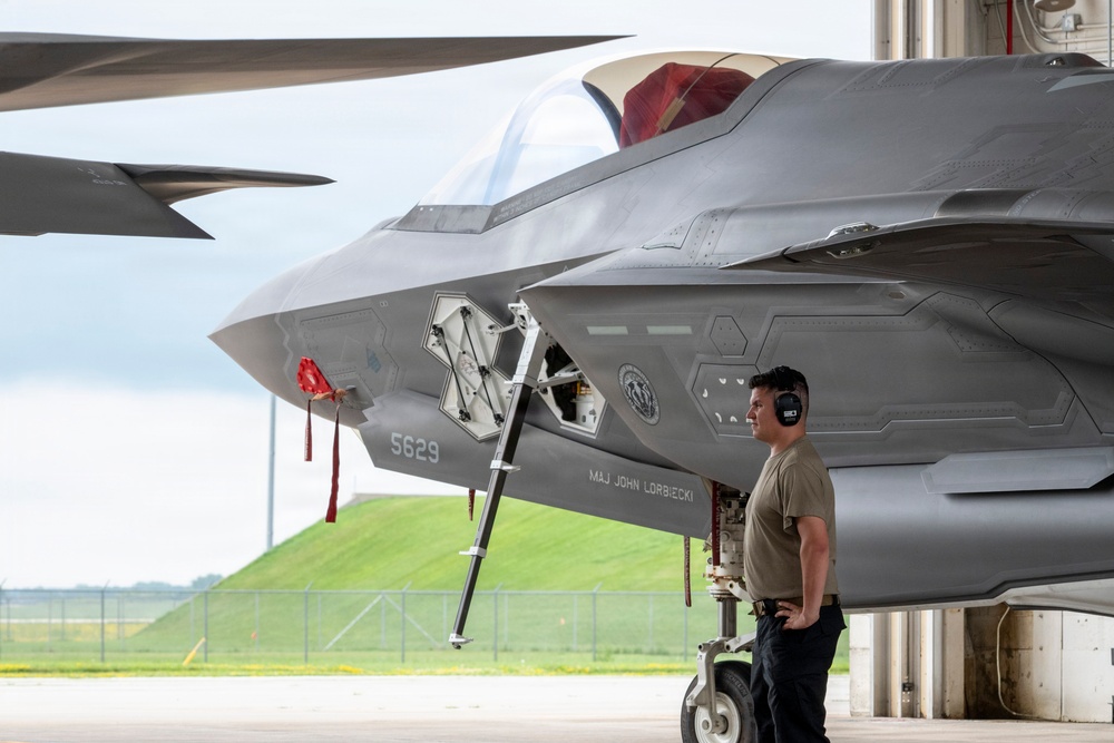 A military ground crew member wearing hearing protection stands beside an F-35 Lightning II stealth fighter jet, identified by tail number 5629 and assigned to 'MAJ JOHN LORBIECKI'. The aircraft is parked on a flight line apron, with a maintenance access panel open on the fuselage. The setting appears to be a military air base with green grass and fencing visible in the background.