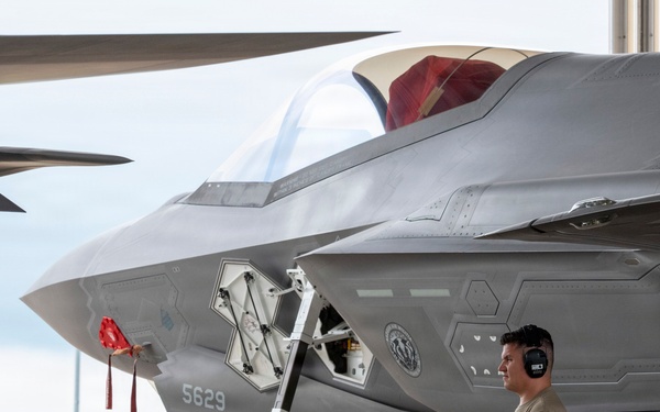 A military ground crew member wearing hearing protection stands beside an F-35 Lightning II stealth fighter jet, identified by tail number 5629 and assigned to 'MAJ JOHN LORBIECKI'. The aircraft is parked on a flight line apron, with a maintenance access panel open on the fuselage. The setting appears to be a military air base with green grass and fencing visible in the background.