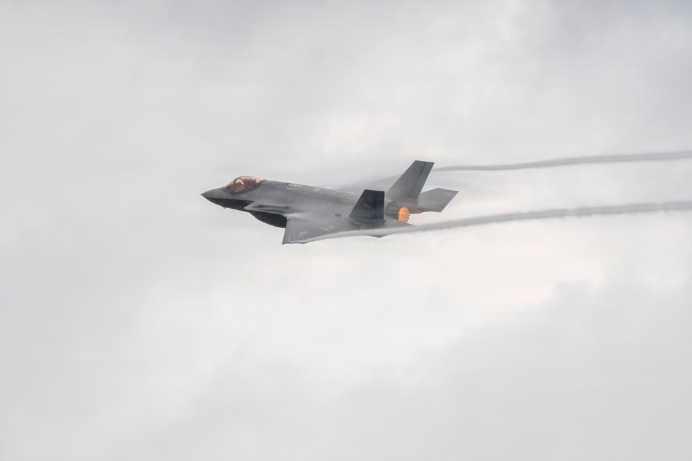 A single-engine stealth fighter jet, consistent with an F-35 Lightning II, is captured in flight against an overcast sky. The aircraft is banking slightly and appears to be at high speed, with visible afterburner flame and condensation vapor trails streaming from the wings and fuselage. The image appears to be taken at an airshow or during a demonstration flight.