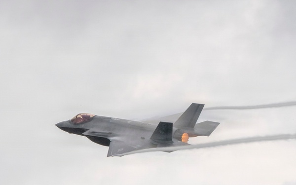A single-engine stealth fighter jet, consistent with an F-35 Lightning II, is captured in flight against an overcast sky. The aircraft is banking slightly and appears to be at high speed, with visible afterburner flame and condensation vapor trails streaming from the wings and fuselage. The image appears to be taken at an airshow or during a demonstration flight.