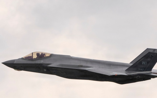 A single-seat stealth fighter jet, identifiable as an F-35A Lightning II, is captured in level flight against an overcast sky. The aircraft bears tail code 'WI' and serial number '15417', indicating it belongs to a Wisconsin Air National Guard unit. A faint vapor trail is visible streaming from the rear of the aircraft.