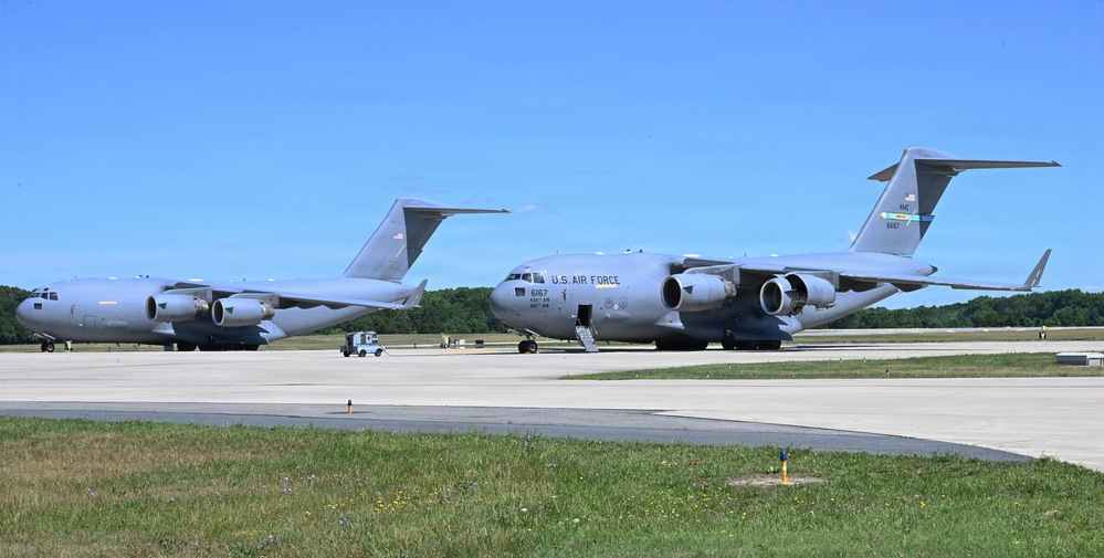 Two U.S. Air Force C-17 Globemaster III cargo aircraft are parked on a flight line tarmac under clear blue skies. The foreground aircraft bears the tail number 6167 and 'U.S. AIR FORCE' markings, with a door open and ground support equipment nearby. A second C-17 is visible in the background to the left.