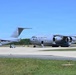 Two U.S. Air Force C-17 Globemaster III cargo aircraft are parked on a flight line tarmac under clear blue skies. The foreground aircraft bears the tail number 6167 and 'U.S. AIR FORCE' markings, with a door open and ground support equipment nearby. A second C-17 is visible in the background to the left.