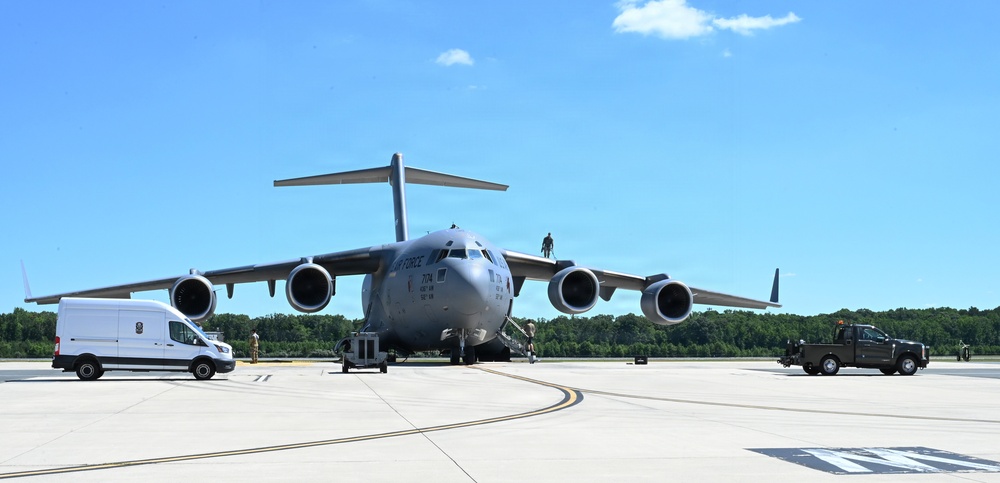 A U.S. Air Force C-17 Globemaster III military cargo aircraft is parked on a tarmac, undergoing ground servicing operations. Multiple ground support vehicles and personnel are visible around the aircraft. The scene appears to be at a military airfield on a clear, sunny day.
