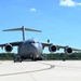 A U.S. Air Force C-17 Globemaster III military cargo aircraft is parked on a tarmac, undergoing ground servicing operations. Multiple ground support vehicles and personnel are visible around the aircraft. The scene appears to be at a military airfield on a clear, sunny day.