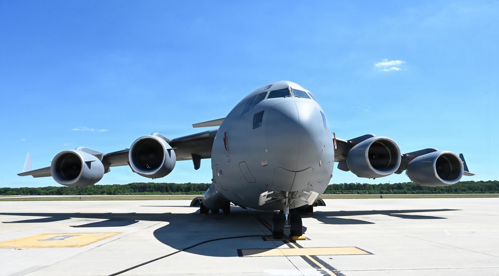 A large military transport aircraft, consistent with a Boeing C-17 Globemaster III, is parked on a concrete tarmac viewed from a direct front-facing perspective. The aircraft is painted in overall gray livery and features four high-bypass turbofan engines mounted under the wings. The setting appears to be a military airfield on a clear, sunny day with green tree lines visible in the background.