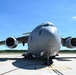 A large military transport aircraft, consistent with a Boeing C-17 Globemaster III, is parked on a concrete tarmac viewed from a direct front-facing perspective. The aircraft is painted in overall gray livery and features four high-bypass turbofan engines mounted under the wings. The setting appears to be a military airfield on a clear, sunny day with green tree lines visible in the background.