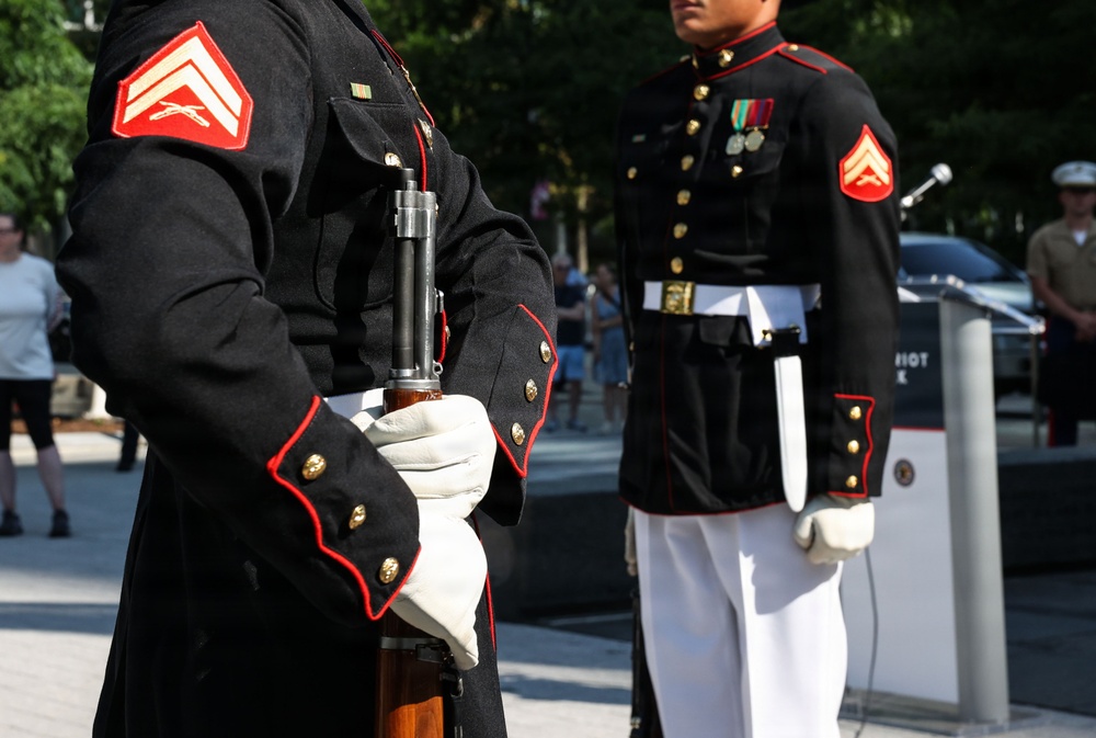 Two United States Marine Corps personnel in dress blue uniforms are standing at attention during what appears to be a formal ceremony or memorial event. Both Marines hold the rank of Corporal, identifiable by the chevron insignia on their sleeves. The foreground Marine is holding a rifle with a fixed bayonet while wearing white gloves, consistent with ceremonial guard duty.