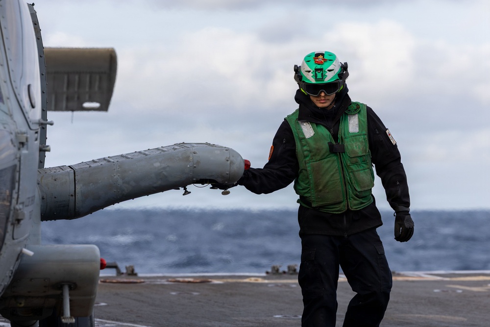 A naval deck crew member wearing a green safety vest and green helmet is handling a helicopter on a ship's flight deck at sea. The personnel appears to be conducting pre-flight or post-flight deck operations, gripping what appears to be a helicopter's tail boom or sensor pod. Overcast skies and open ocean are visible in the background, suggesting active maritime operations.