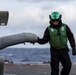A naval deck crew member wearing a green safety vest and green helmet is handling a helicopter on a ship's flight deck at sea. The personnel appears to be conducting pre-flight or post-flight deck operations, gripping what appears to be a helicopter's tail boom or sensor pod. Overcast skies and open ocean are visible in the background, suggesting active maritime operations.