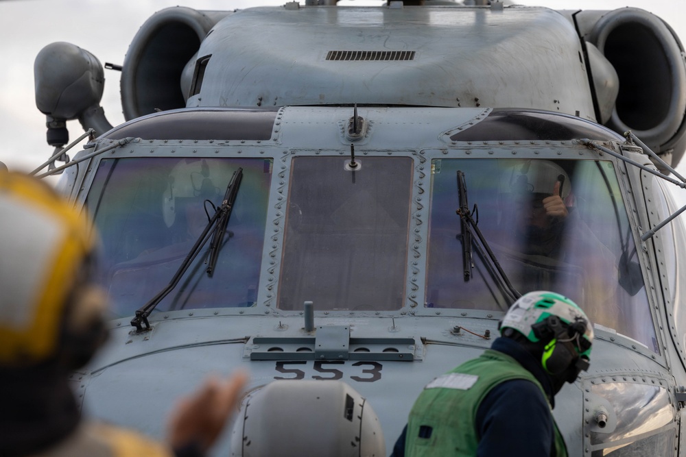 A close-up view of the nose and cockpit section of a military helicopter marked with the number '553'. Ground crew personnel wearing colored helmets and vests are visible in the foreground, and a pilot or crew member inside the cockpit appears to be giving a thumbs-up gesture. The scene appears to be taking place on a naval vessel flight deck during pre-flight or post-flight operations.