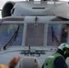 A close-up view of the nose and cockpit section of a military helicopter marked with the number '553'. Ground crew personnel wearing colored helmets and vests are visible in the foreground, and a pilot or crew member inside the cockpit appears to be giving a thumbs-up gesture. The scene appears to be taking place on a naval vessel flight deck during pre-flight or post-flight operations.