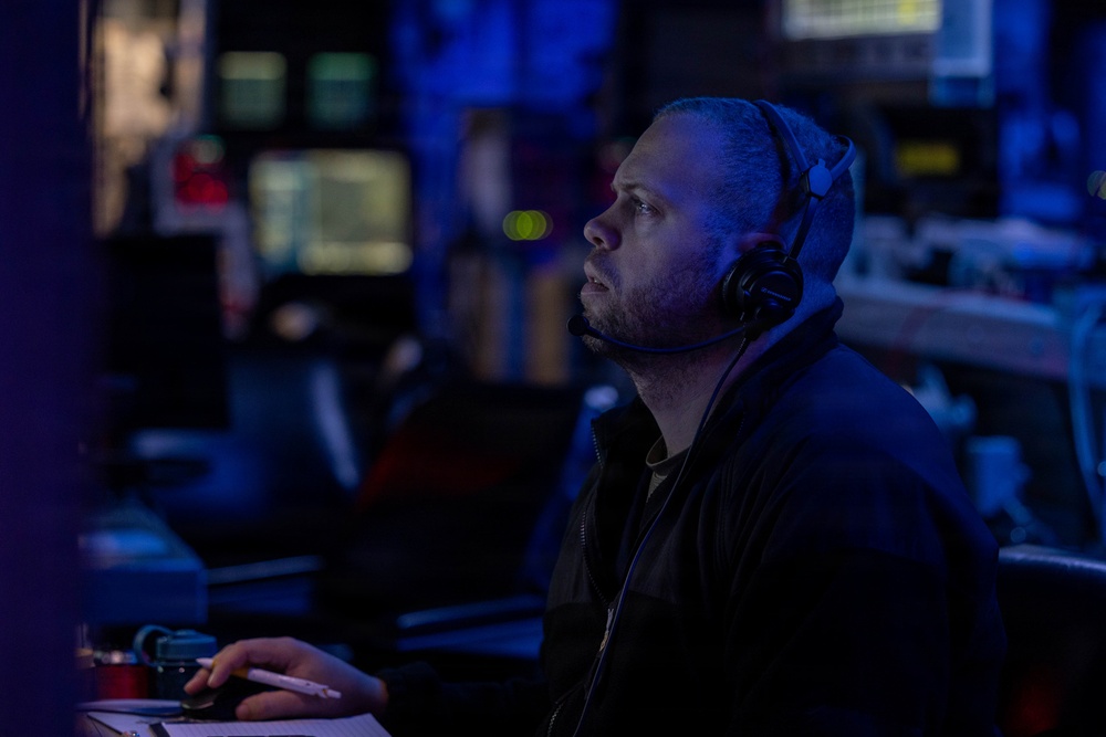 A man wearing a communications headset with a boom microphone is seated at a control station in a dimly lit operations room illuminated by blue ambient lighting. He appears to be monitoring screens or displays, looking upward and to the right with focused attention. The environment suggests a military combat information center, naval operations room, or similar command and control facility.