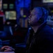 A man wearing a communications headset with a boom microphone is seated at a control station in a dimly lit operations room illuminated by blue ambient lighting. He appears to be monitoring screens or displays, looking upward and to the right with focused attention. The environment suggests a military combat information center, naval operations room, or similar command and control facility.