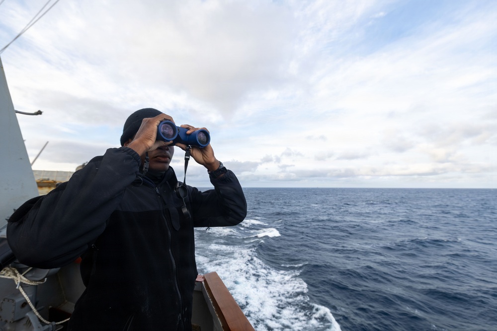 A person dressed in black cold-weather gear and a black beanie stands on the deck of a moving vessel, scanning the open ocean through blue binoculars. The ship's wake is visible in the background, indicating the vessel is underway. The overcast sky and choppy sea suggest cool, maritime conditions.