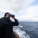 A person dressed in black cold-weather gear and a black beanie stands on the deck of a moving vessel, scanning the open ocean through blue binoculars. The ship's wake is visible in the background, indicating the vessel is underway. The overcast sky and choppy sea suggest cool, maritime conditions.