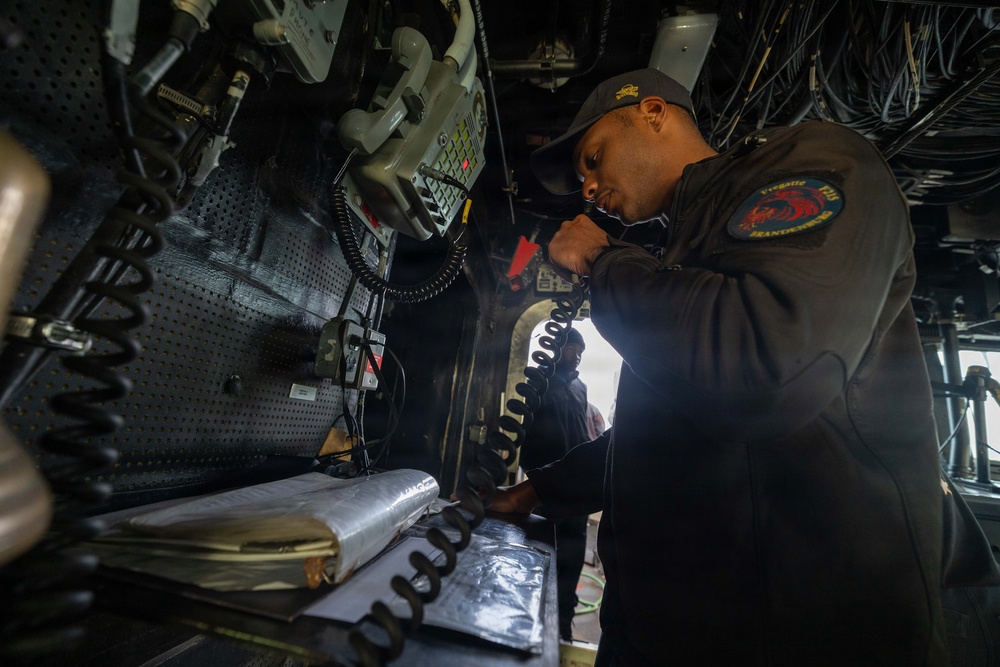 A naval service member in dark coveralls is using a coiled-cord radio handset in what appears to be a ship's bridge or combat information center. The individual is wearing a unit patch on their left sleeve and a dark baseball cap with an emblem. The environment is densely packed with electronic equipment, cables, and communication systems typical of a military vessel.