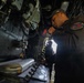 A naval service member in dark coveralls is using a coiled-cord radio handset in what appears to be a ship's bridge or combat information center. The individual is wearing a unit patch on their left sleeve and a dark baseball cap with an emblem. The environment is densely packed with electronic equipment, cables, and communication systems typical of a military vessel.