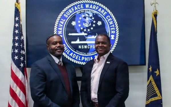 Two men in business attire are posing together in front of a Naval Surface Warfare Center Philadelphia Division (NSWCPD) logo displayed on a screen. The setting appears to be an official government or military facility. An American flag and what appears to be a state flag flank the individuals on either side.